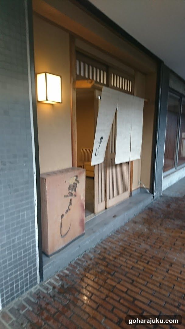 A traditional-style shop entrance with wooden sliding doors and a noren curtain hanging at the doorway. A square wall-mounted light fixture illuminates the entrance, and a large rectangular signboard is placed on the ground near the door. The building features tiled walls and a brick-paved walkway leading to the entrance.