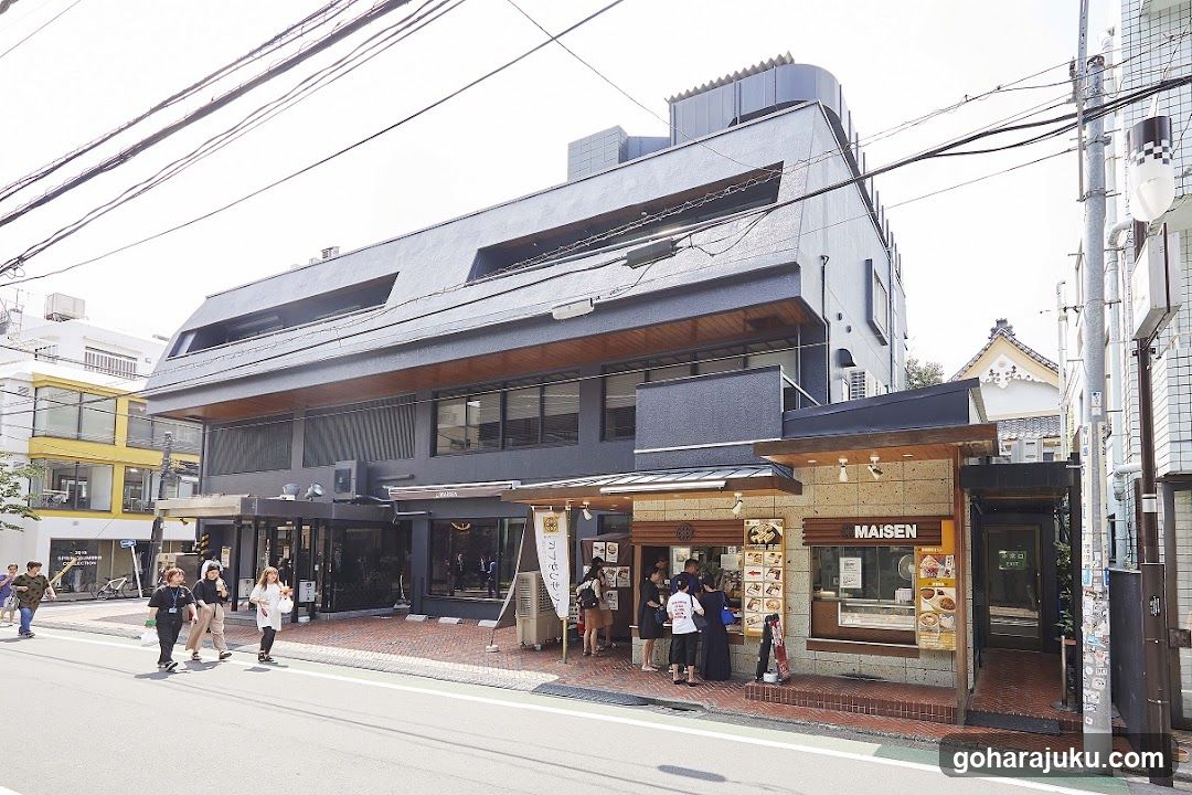 A modern two-story building with a dark facade and wooden accents, featuring a ground-floor entrance with people walking by. A smaller attached structure displays menu boards and signage.