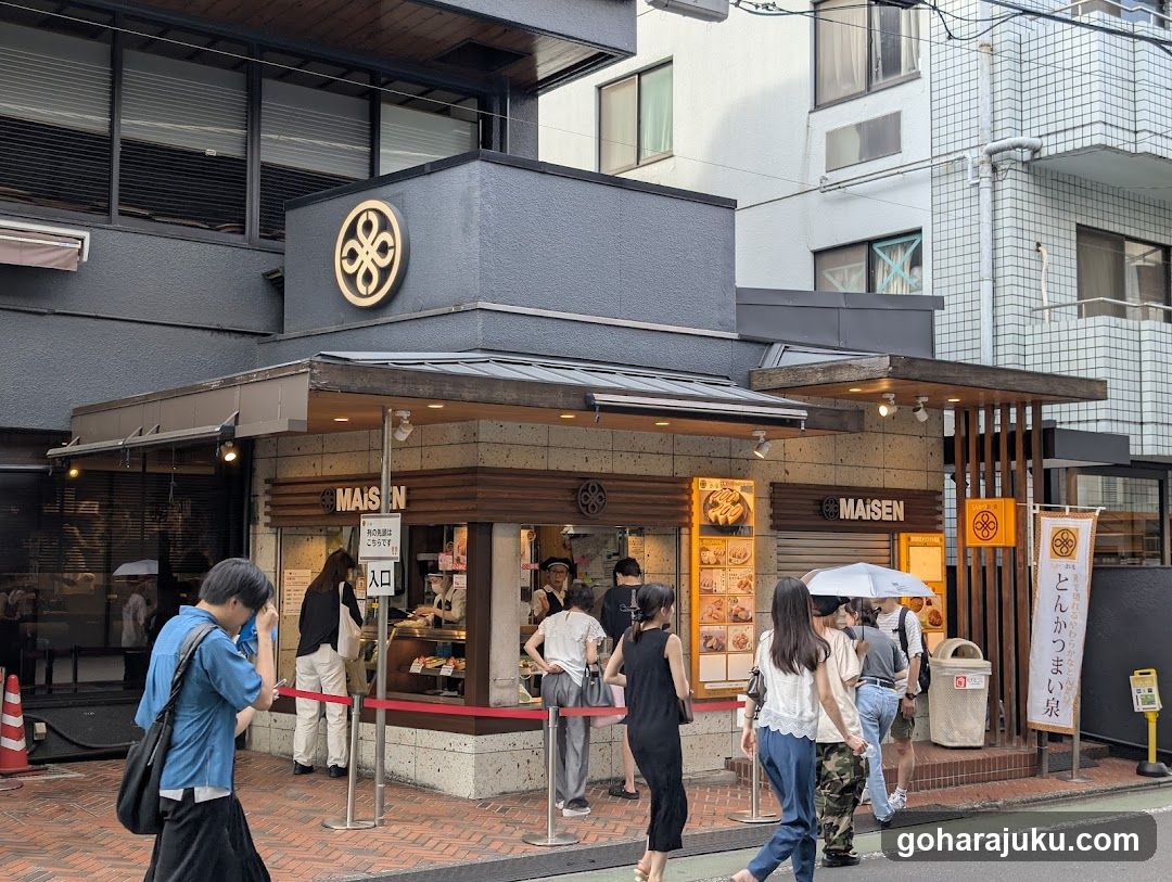 A modern shop front with a dark gray facade and wooden accents, featuring the 'MAISON' sign on both sides of the entrance. A queue of customers is lined up behind red ropes, waiting to enter. The building has large windows displaying food items inside, and there are vertical banners with Japanese text on the right side.