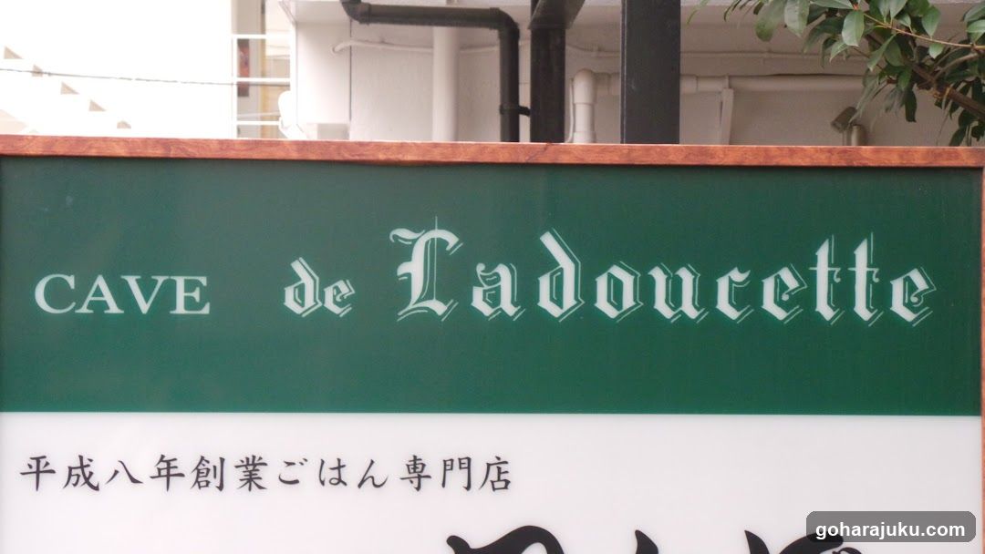 A close-up photo of a shop sign on the exterior facade. The green background features white text reading 'CAVE de Ladoucette' in stylized fonts. Below, Japanese characters indicate it is a specialty restaurant established in Heisei Year 8 (1996). No menu or interior view visible.