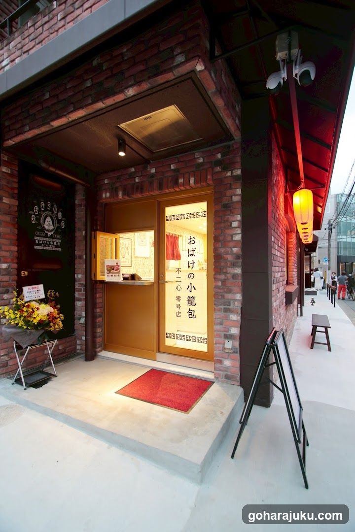A brick building entrance with a wooden door and window displaying Japanese text 'おばけの小籠包' (Oni's dumplings). A red lantern hangs outside, and a floral arrangement sits near the entrance.