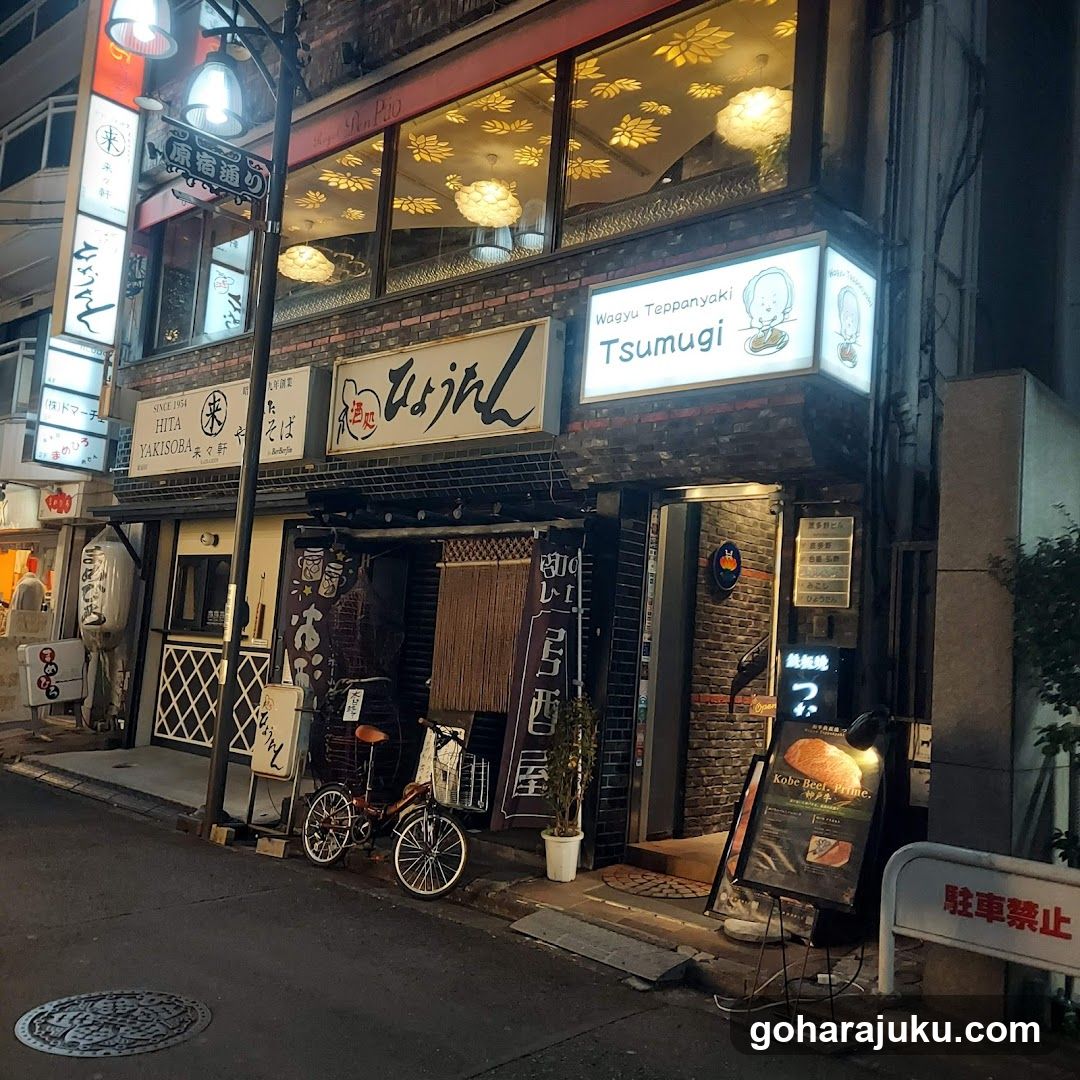 Night view of a traditional Japanese restaurant facade with illuminated signage reading 'Wagyu Teppanyaki Tsumugi'. Brick walls, wooden doors, hanging lanterns, and bicycles parked outside.