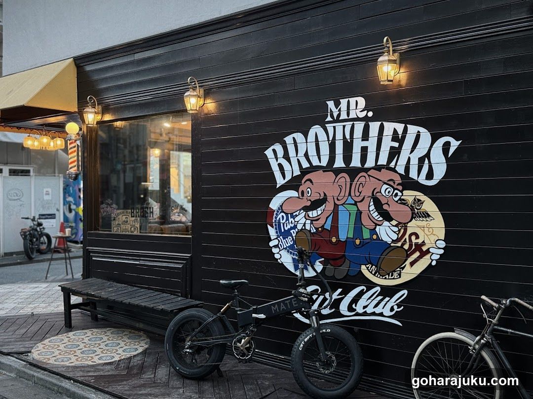 A black wooden storefront with a large mural of two cartoon characters holding beer mugs and the text 'MR. BROTHERS Bar Club'. Two vintage-style lantern lights are mounted on the wall above the mural. A bicycle is parked in front, and another bike is partially visible to the right. A bench sits near the entrance, which has a glass door and window displaying interior items.