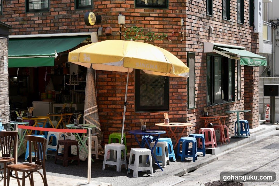 A brick building exterior featuring a cafe entrance with green awnings and colorful plastic stools under yellow umbrellas. Visible signage includes 'SINGHA' on the umbrella and partial neon lettering above.