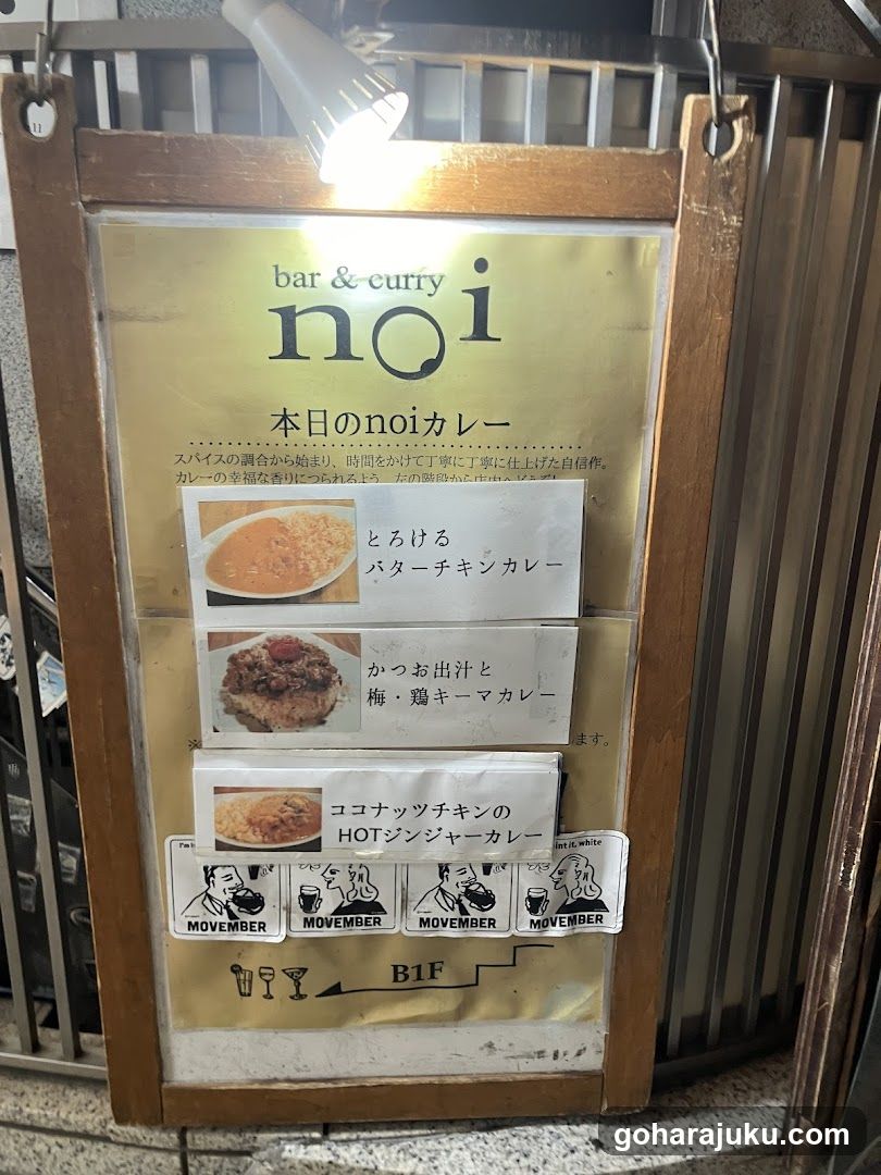 A wooden-framed menu board is mounted on the exterior of a shop, illuminated by an overhead light. The sign features Japanese and English text listing daily curry options.