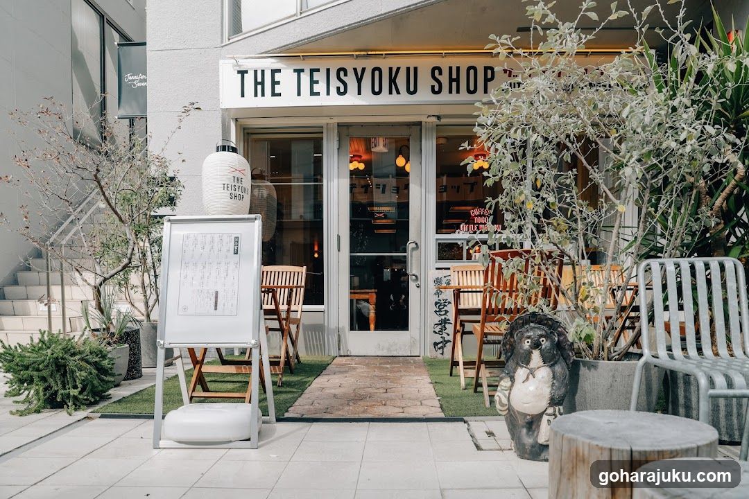 A modern storefront with a white facade and large glass doors displaying warm interior lighting. A prominent sign reads 'THE TEISYOKU SHOP'. Outdoor seating includes wooden chairs, metal chairs, and potted plants. A freestanding menu board is positioned to the left of the entrance.