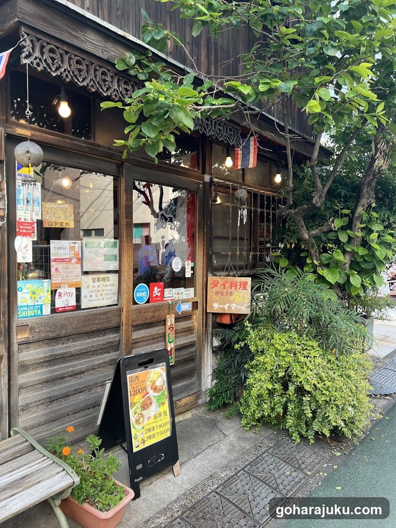 A wooden shop front with ornate carvings, glass doors adorned with posters and signs, hanging lanterns, and lush greenery surrounding the entrance.