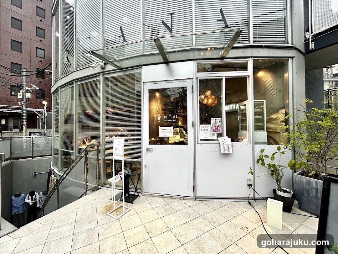 The photo shows the exterior entrance of a modern coffee shop with glass walls and white doors. The word 'INITIAL' is visible on the window, along with smaller text below it. There are several signs posted near the entrance, including one that appears to be a menu or price list. Inside, warm lighting illuminates the interior space, which includes hanging lights and tables.