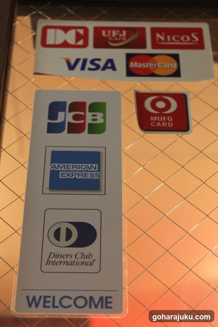A close-up view of a storefront entrance featuring multiple credit card logos affixed to the glass door or window. The visible brands include DC, UFJ GM, NicoS, VISA, MasterCard, JCB, MUFG CARD, American Express, Diners Club International, and a WELCOME sign at the bottom. The background shows a tiled floor with diagonal grout lines.