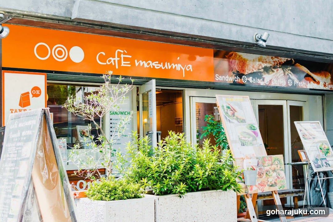 The image shows the storefront of Café Masumiya, featuring an orange sign with white lettering and circular logos. The entrance is flanked by green plants in concrete planters. Menu boards are displayed outside, showcasing sandwiches, salads, coffee, fresh juices, and sweets. A security camera is mounted above the entrance.