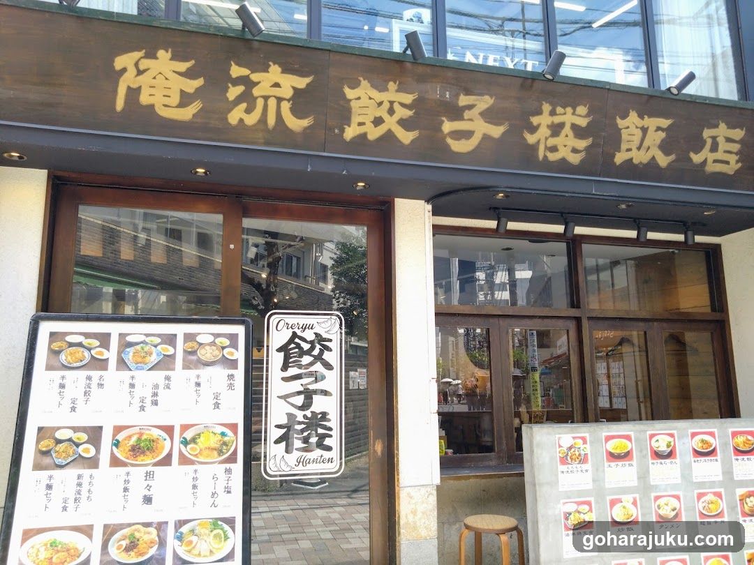 A shop front with a wooden signboard displaying Japanese characters, likely indicating the name of the establishment. The entrance features glass doors and windows, allowing a glimpse into the interior. A menu board is visible outside, showcasing various food items.