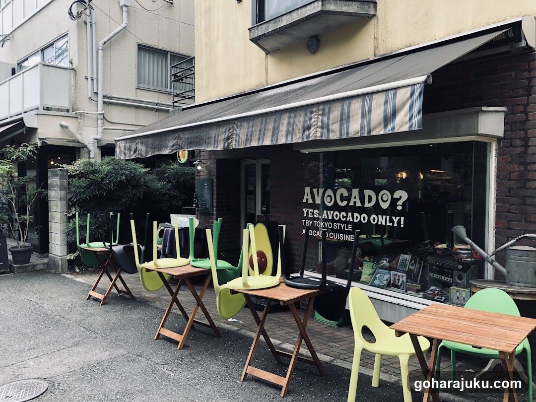 A street-level view of a small cafe or shop with an awning and outdoor seating. The storefront has large glass windows displaying various items, including framed pictures and possibly food products. Several wooden tables and chairs are arranged outside on the sidewalk.
