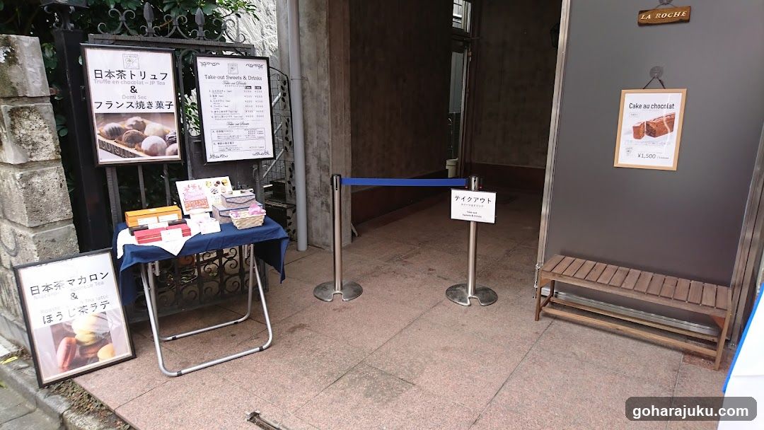 Exterior view of a shop entrance with signage and promotional materials. Visible signs include '日本茶トリュフ & フランス焼き菓子' (Japanese Tea Truffles & French Baked Goods) and 'Cake au chocolat'. A blue rope barrier separates the entrance area, and a wooden bench is placed near the door.