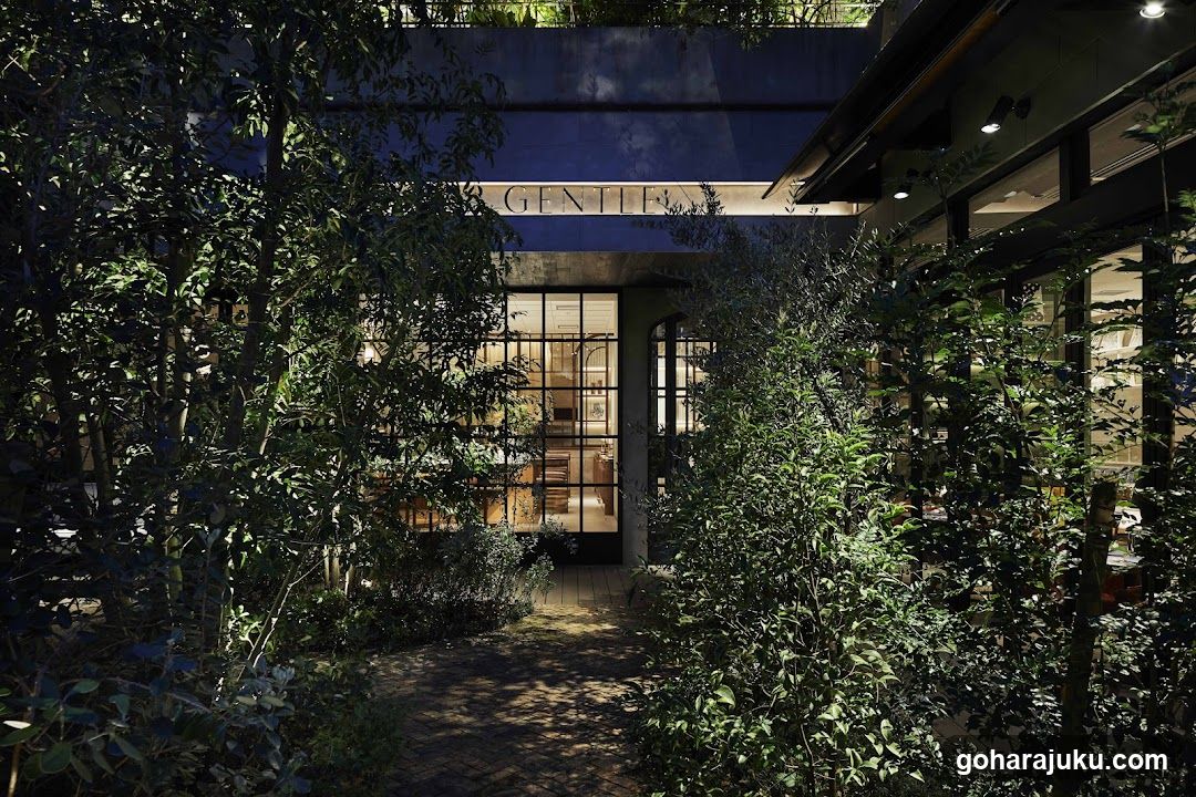 A dimly lit storefront entrance framed by lush greenery and trees, with the word 'GENTLEMEN' visible on a sign above the glass doors.