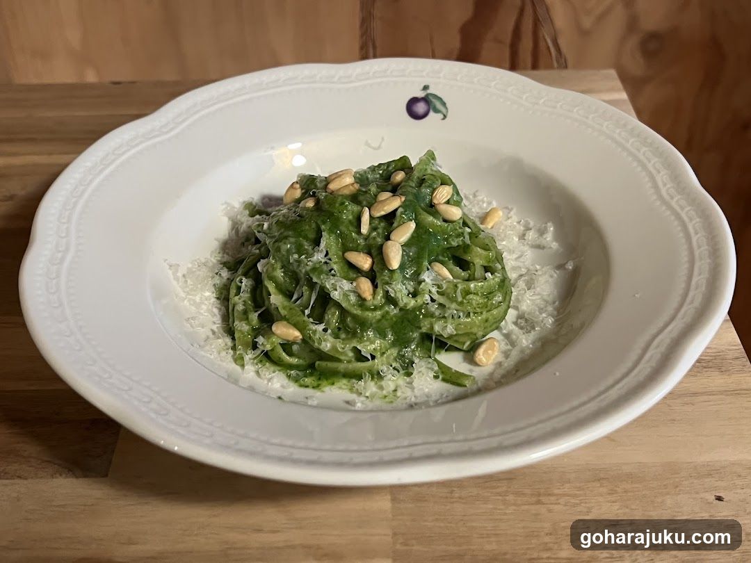 A white bowl containing green pasta, likely pesto-based, topped with pine nuts and grated cheese, set on a wooden table.