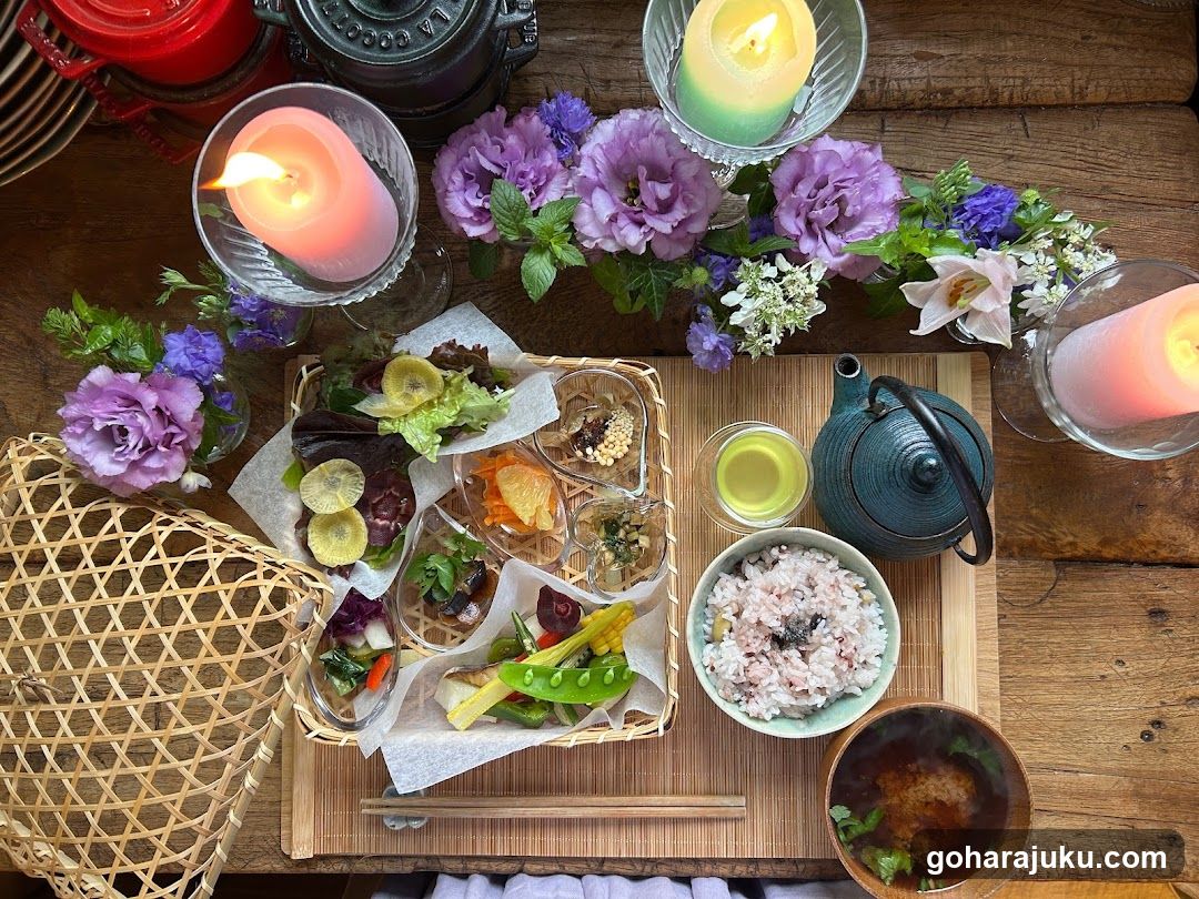 Top-down view of a wooden table set with a woven basket containing assorted appetizers, including pickled vegetables and garnishes. A bowl of rice sits beside a small teapot and tea cup. Decorative elements include lit candles in glass holders, purple flowers, and greenery arranged around the food.