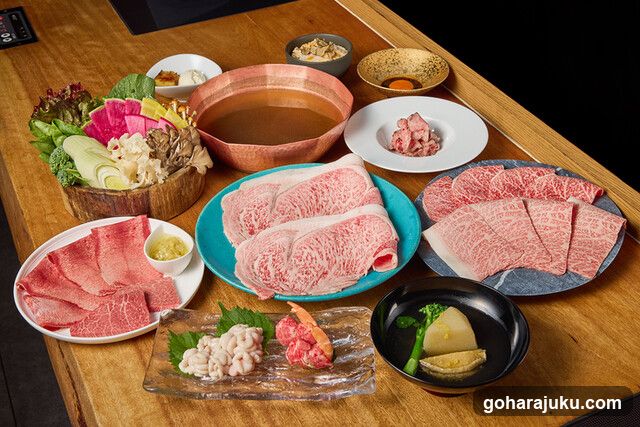 A table set with a variety of Japanese hot pot ingredients, including raw beef slices on plates, vegetables in a wooden bowl, dipping sauces, and small bowls of condiments.