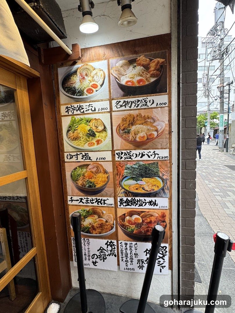 A photo of a ramen shop's exterior entrance, featuring a large menu board on the wall displaying various noodle dishes with prices in yen. The shop has wooden framing and glass doors, with street lights visible above.