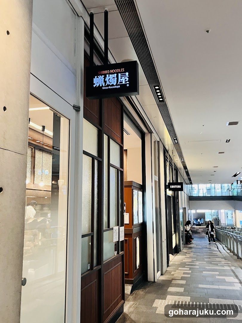 The image shows the exterior entrance of a shop named 'Shinri Noodles' with its signboard prominently displayed above the door. The storefront features glass windows and wooden accents, giving it a modern yet traditional appearance. Inside, there are tables and chairs visible, suggesting a dining area.