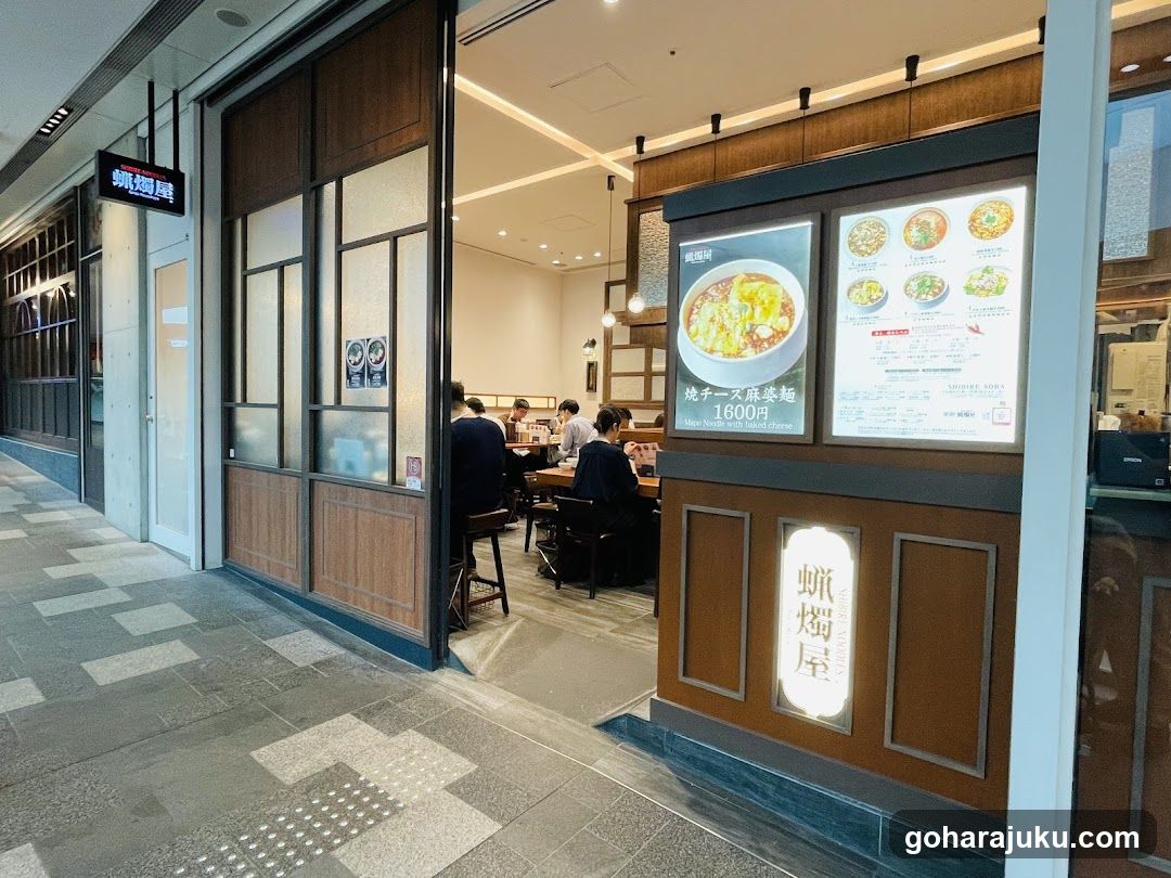 The image shows the exterior entrance of a ramen shop with wooden paneling and glass doors. A lit sign on the wall reads 'NARUTAYA' in vertical Japanese characters. Inside, customers are seated at tables, and staff members are visible near the counter. The interior is warmly lit with hanging pendant lights.