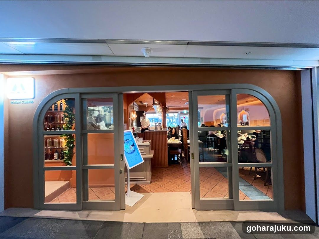 Exterior view of a restaurant with arched glass doors and warm interior lighting. A blue menu stand is placed at the entrance, displaying food items and prices. Inside, diners are seated at tables, and staff members are visible near the counter.