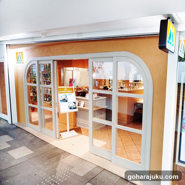 The image shows the exterior entrance of a restaurant or café, featuring large arched glass doors and windows framed in white. The interior is warmly lit, revealing tables, chairs, and kitchen equipment visible through the glass. A menu stand is placed outside near the entrance, displaying food items and prices. The building has a beige facade with signage above the door.