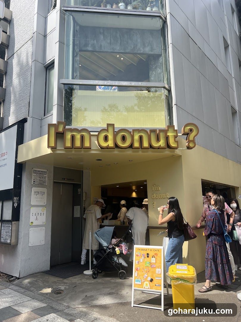 Photo of the exterior entrance and facade of a shop with 'I'm donut?' signage mounted on the building. Customers are gathered at the counter, some holding drinks or waiting in line. A yellow trash bin is visible near the entrance.