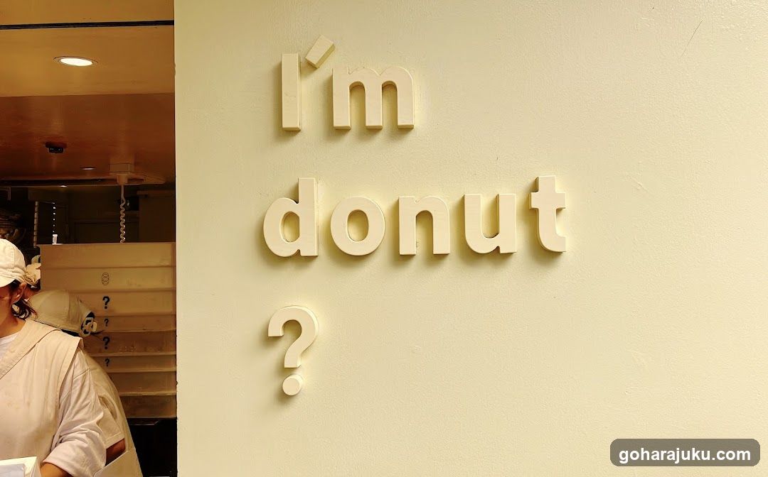 A cream-colored wall features raised, light-colored lettering spelling 'I'm donut?' with a question mark below. To the left, part of an interior is visible, including a person in white attire and wooden shelving units.