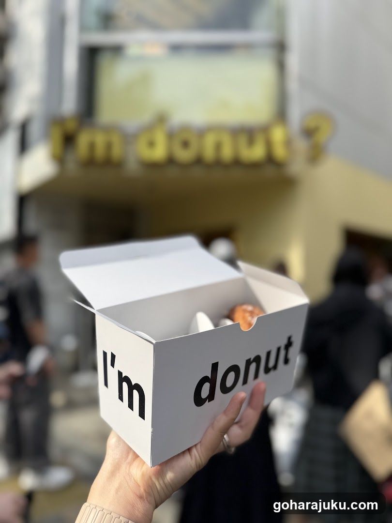 A person holds a white box labeled 'I'm donut' in the foreground, with a blurred shop sign reading 'I'm donut?' visible on the building facade behind.