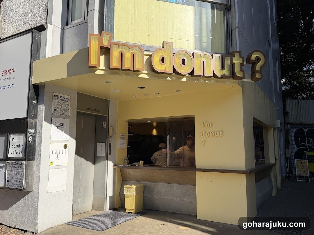 A yellow storefront with large 3D signage reading 'I'm donut?' above the entrance. A service window is visible on the right side, showing staff inside. A small yellow trash bin sits near the door. Posters and notices are affixed to the left wall.