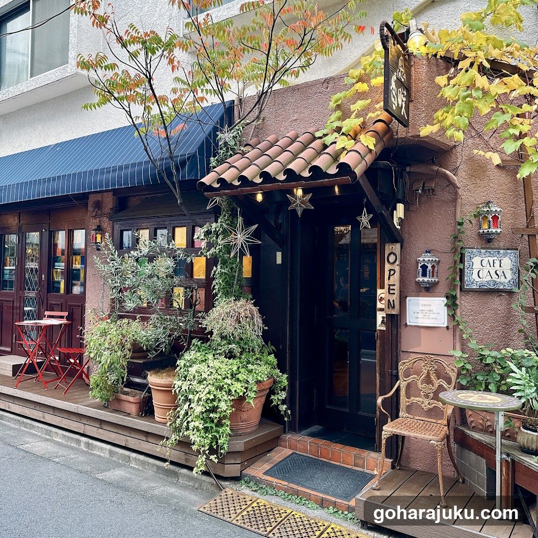 A charming cafe entrance with terracotta roof tiles, potted plants, and a small outdoor seating area. The building features warm-toned walls, decorative lanterns, and an open door sign.