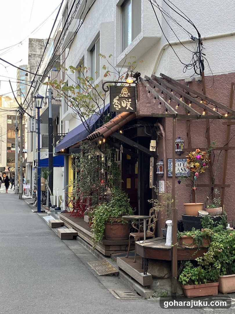 A narrow urban street scene featuring a small shop or cafe entrance on the right side of the frame. The building has white walls with exposed wiring overhead, and a wooden pergola structure adorned with string lights extends over part of the facade. A dark signboard hangs above the doorway displaying Japanese characters (likely indicating 'flavor' or similar). Outside, there are potted plants in terracotta pots arranged along the sidewalk and near the entrance. Red folding chairs and tables suggest an outdoor dining area. To the left, other storefronts line the street, including one with blue awninging. Pedestrians walk further down the road under daylight conditions.