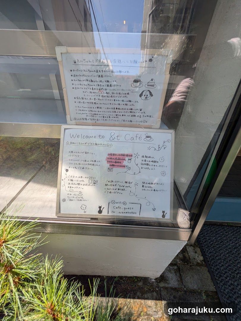 Photo of a cafe entrance with two whiteboards mounted on the glass door. The top board contains Japanese text and illustrations of coffee cups. The bottom board says 'Welcome to Cafe Guest' in English, followed by Japanese text and small doodles including cats and flowers.