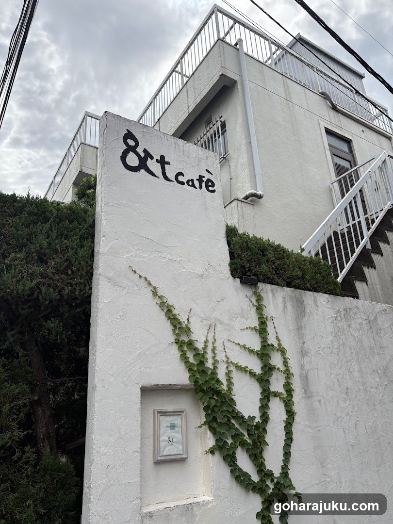 White stucco building facade with black handwritten-style sign reading '&t cafe' on upper left corner. Green ivy climbs down the right side of the wall. A small framed notice is embedded in the lower left portion. White metal railing staircase ascends to second floor on right. Overhead power lines visible against cloudy sky.