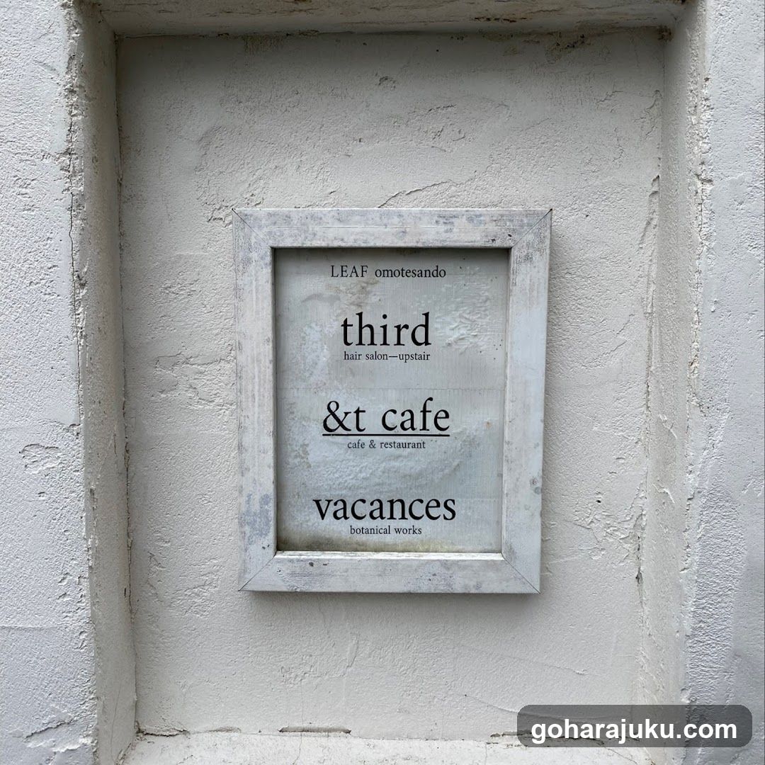 A framed sign mounted on a textured white wall, displaying the names of three businesses: 'third hair salon—upstairs', '&t cafe (cafe & restaurant)', and 'vacances botanical works'. The frame is rustic and weathered.