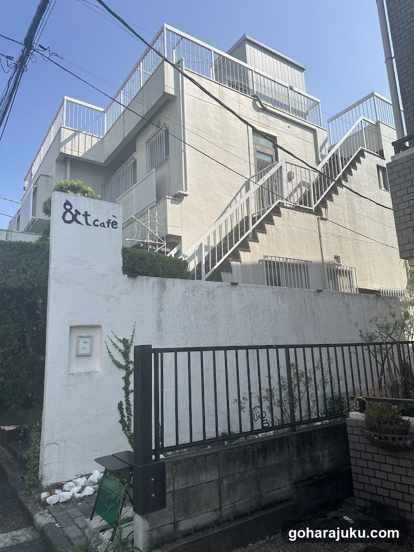 A multi-story building with a white facade and metal railings, featuring an external staircase leading to upper levels. A prominent sign on the wall reads '8&1 cafe'. The entrance is partially obscured by a black metal fence.