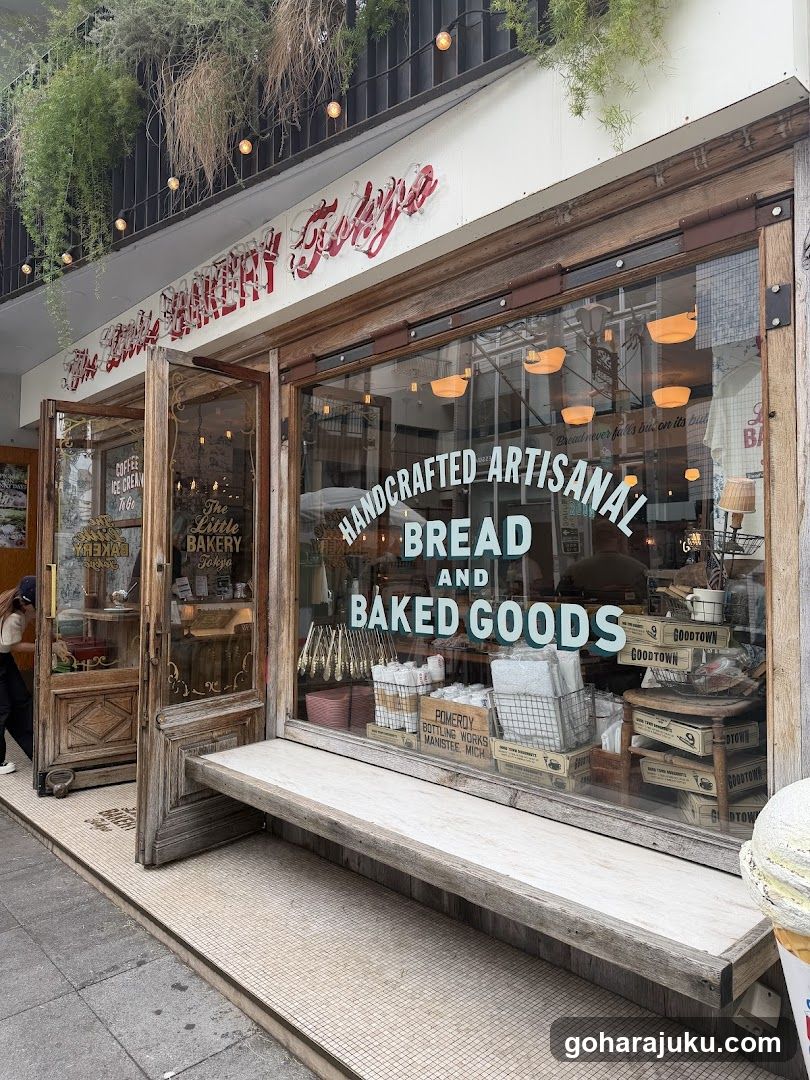 A photo of the physical storefront facade featuring a wooden entrance and large glass windows displaying baked goods. Visible signage includes 'The Little Bakery Tokyo' in red script above the door, and 'HANDCRAFTED ARTISANAL BREAD AND BAKED GOODS' on the window. Hanging plants adorn the upper balcony area.
