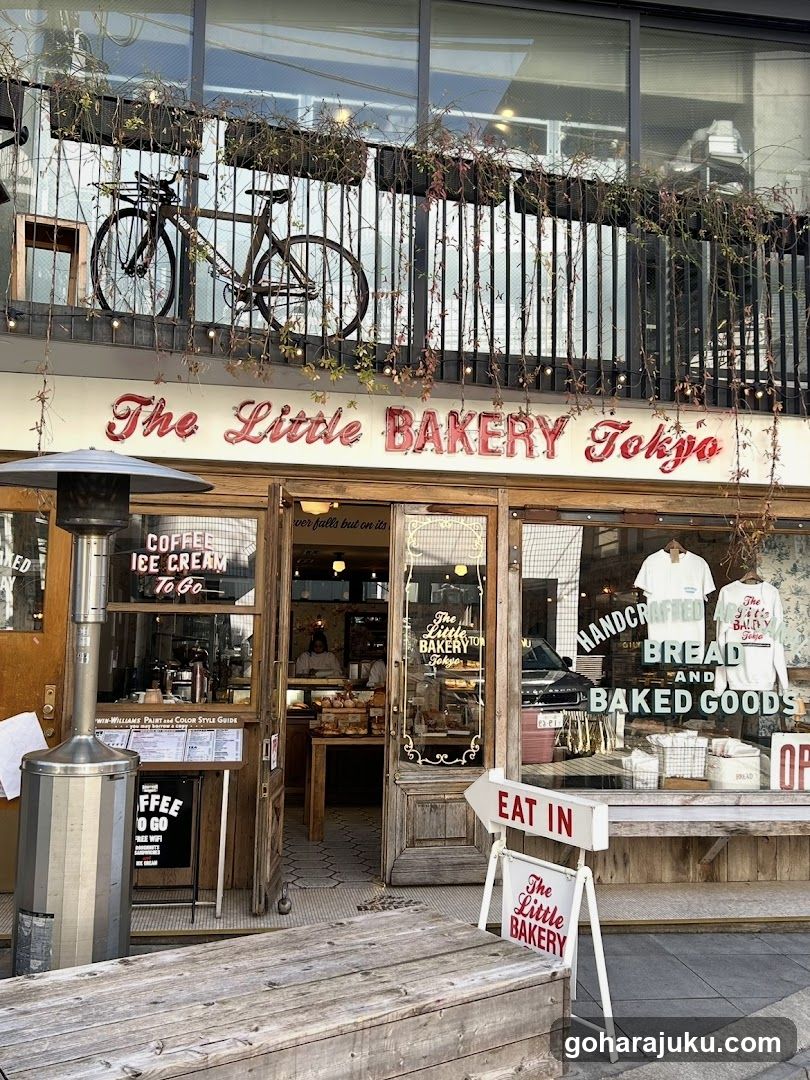 A charming bakery storefront with a sign reading 'The Little BAKERY Tokyo' in red cursive and block letters. A bicycle is mounted on the upper balcony, adorned with trailing plants. The entrance features wooden doors and windows displaying baked goods and merchandise like t-shirts. An outdoor heater stands near the entrance, and a menu board lists coffee and ice cream options.