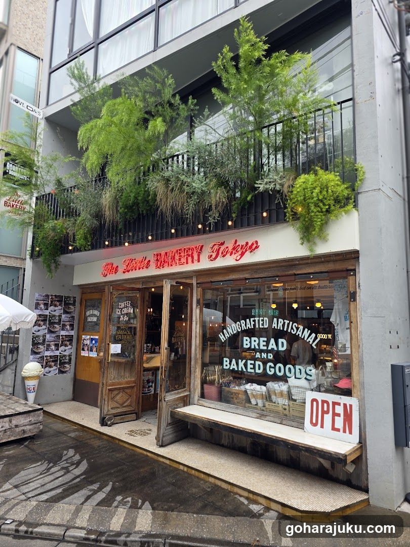 A street-level view of The Little Bakery Tokyo storefront, featuring a wooden entrance with glass doors and large display windows showcasing baked goods. Above the entrance is a balcony adorned with lush greenery and hanging plants. A red neon sign reads 'The Little BAKERY Tokyo' above the shop front. Inside, shelves are visible with bread and other items on display. An open sign sits near the entrance.