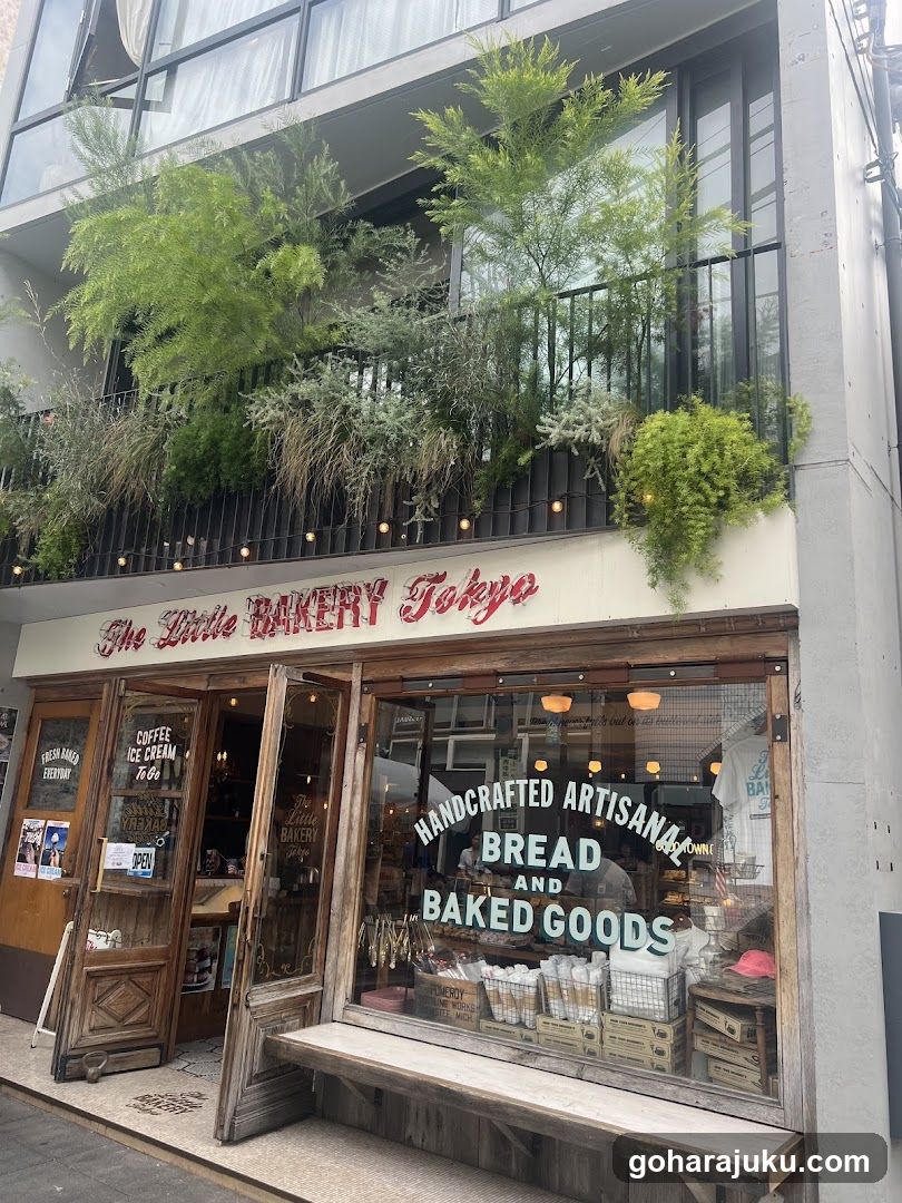 A charming bakery storefront with a wooden facade and large glass windows displaying baked goods. The sign reads 'The Little BAKERY Tokyo' in red cursive letters above the entrance. Inside, shelves are stocked with breads and pastries, while hanging plants adorn the balcony above.
