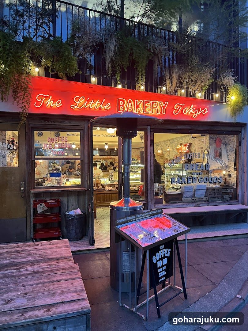 A brightly lit storefront with a red neon sign reading 'The Little BAKERY Tokyo'. The entrance features glass doors and windows displaying baked goods inside. A menu board stands outside, listing coffee options like 'COFFEE TO GO' and other items.