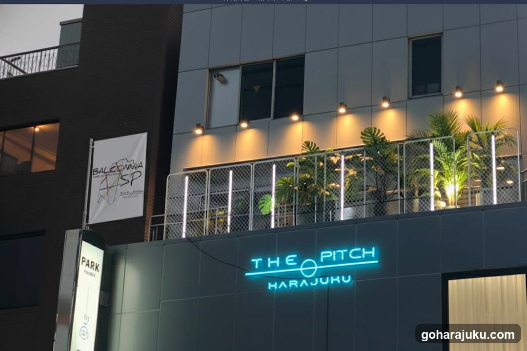 Evening shot of a modern building facade with illuminated signage reading 'THE PITCH HARAJUKU' in neon blue letters on the lower section. Above, a balcony area features potted plants and vertical lighting strips along the railing. A white banner to the left displays 'BALCONY SPA'. The scene is lit by warm spotlights against a dark sky.