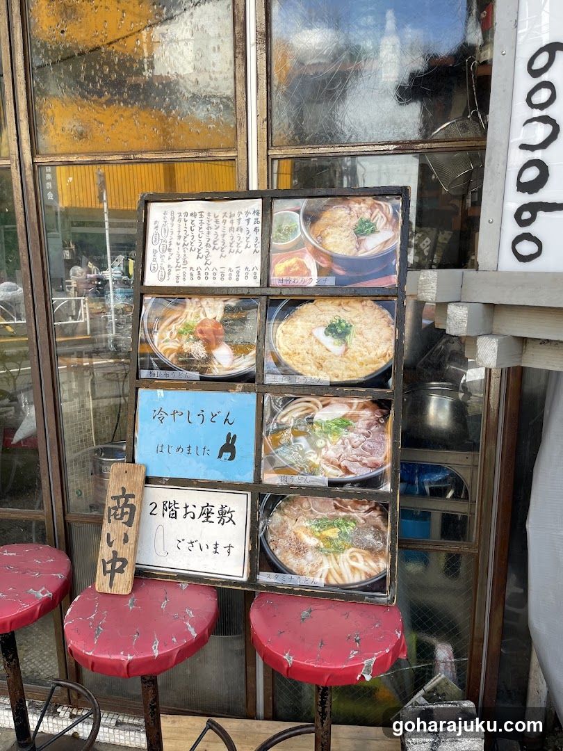 A photo of a shop's exterior entrance, featuring a large window display showcasing various ramen dishes and a sign indicating 'Closed' (商い中). The menu board is placed outside the store, displaying images of different noodle soups.