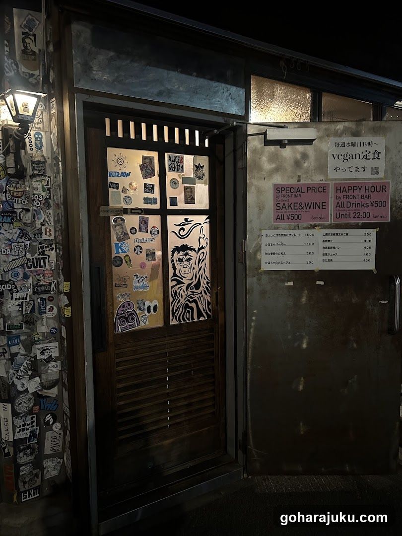 A dimly lit storefront with a wooden door covered in stickers and artwork, including a zebra-striped face. A sign on the right advertises vegan dining hours and happy hour specials.