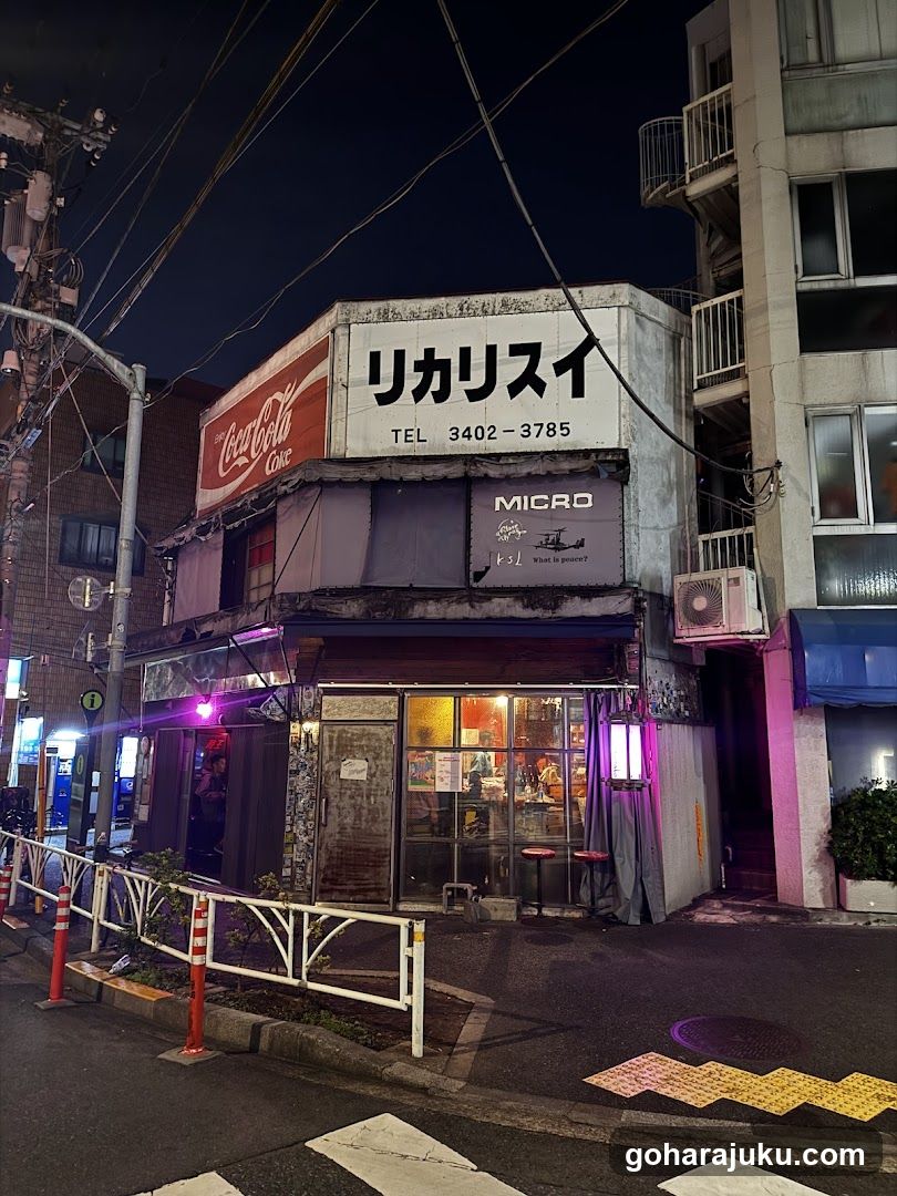 A nighttime photo of a small shop on a city street corner. The building has a white sign with Japanese characters and phone number 'TEL 3402-3785'. Below, there is a Coca-Cola logo on the left side. A purple neon light illuminates the entrance area. There are glass doors leading into the store, which appears to have warm interior lighting visible through the windows. An air conditioning unit is mounted on the wall next to the entrance. Power lines crisscross above against a dark sky.