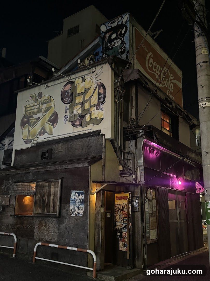 A dimly lit shop at night, featuring a large graffiti-style sign on the upper facade and purple neon lighting above the entrance. The building has a rustic wooden exterior with visible posters and flyers near the door.