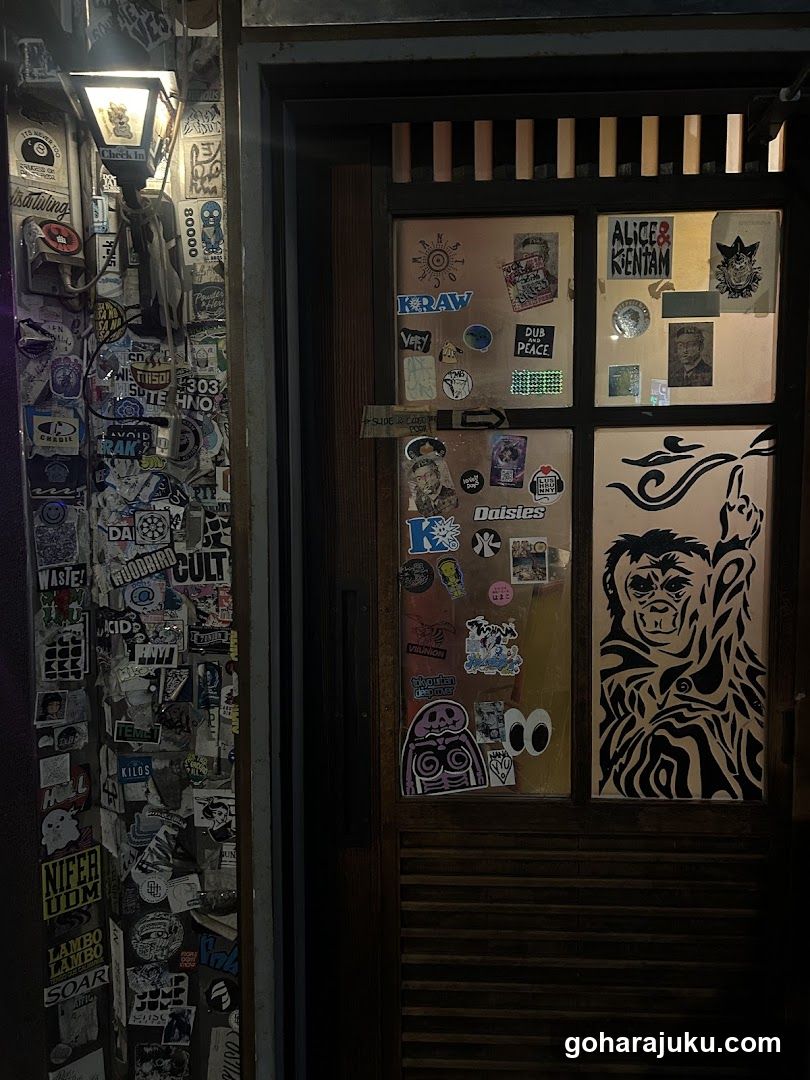 A dimly lit exterior view of a shop entrance featuring glass doors and side panels heavily covered in stickers, including brand logos like KRAW, Alice & Kentan, and Daisies. A stylized monkey illustration is visible on the right door panel. The left wall displays additional sticker clusters under an illuminated lantern-style light fixture.