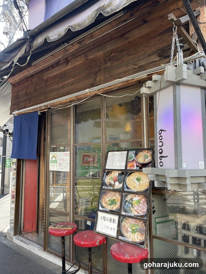 A wooden shop front with a blue noren curtain, glass windows displaying interior decor, and red stools outside. A menu board stands in front showing ramen dishes.