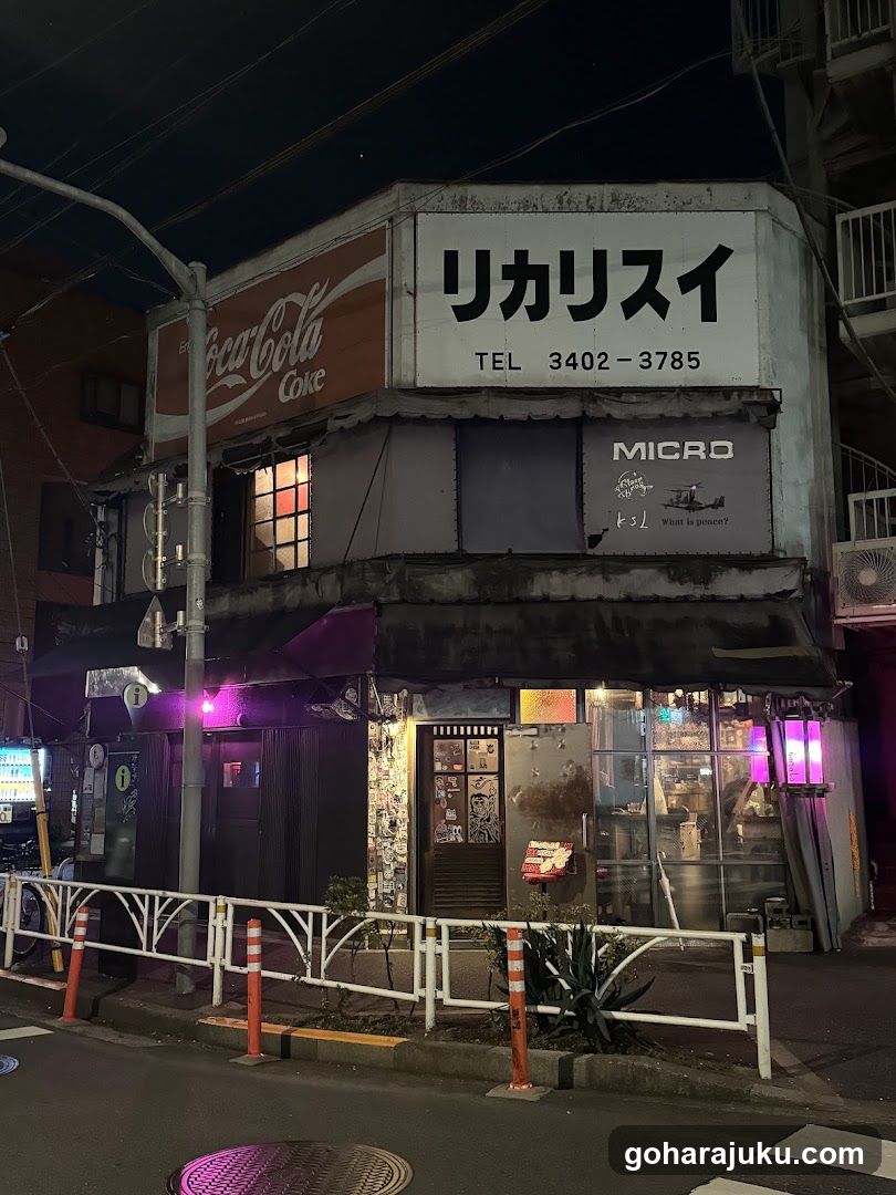 A corner building at night featuring a Coca-Cola sign on the left, a large white sign with Japanese characters (リカリスイ) and phone number above the entrance, and a 'MICRO' advertisement to the right. The shop has glass doors and windows, purple lighting accents, and is surrounded by a low fence.