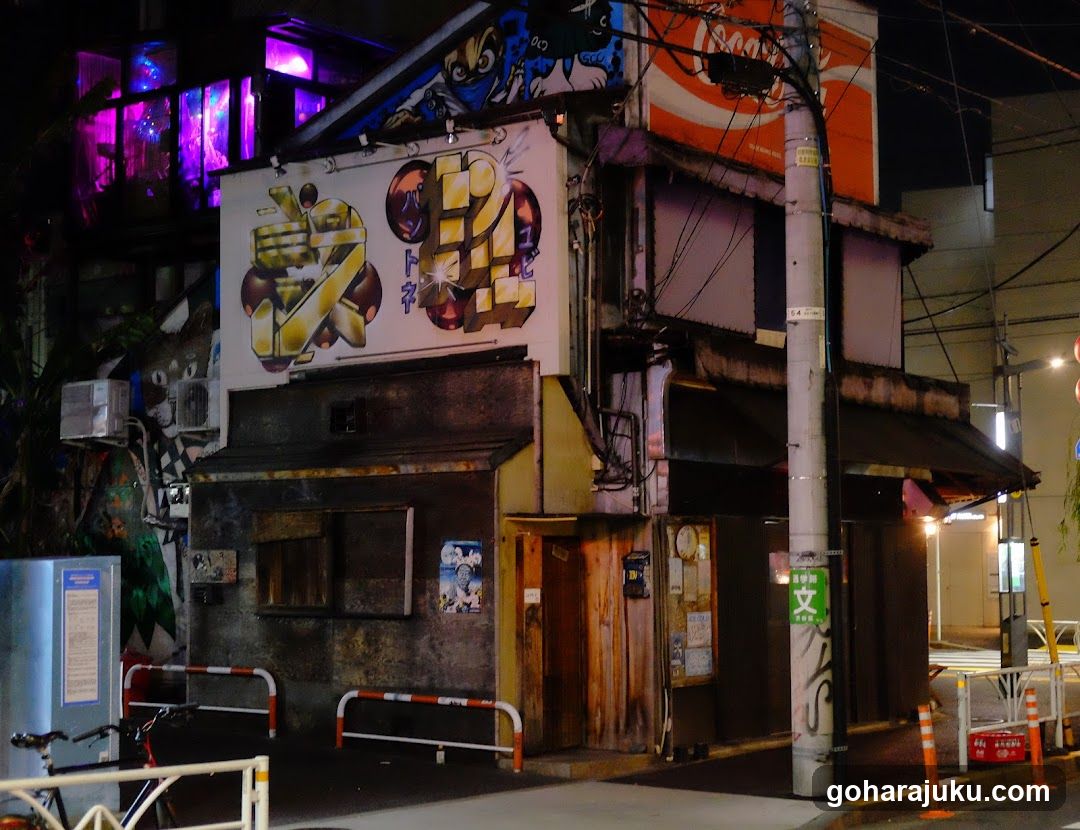 A dimly lit street corner at night featuring a building adorned with vibrant graffiti art on its facade. The structure has multiple levels, with visible signage including a large illuminated sign above the entrance displaying stylized characters and graphics. A smaller vertical sign is mounted near the ground level. The scene includes urban elements such as utility poles, wires, and traffic barriers.
